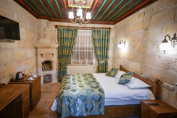 A bedroom with a bed, wooden headboard, a nightstand, a bedside table, a fireplace, and stone walls. - Cappadocia Sightseeing Hotel (Nevsehir)