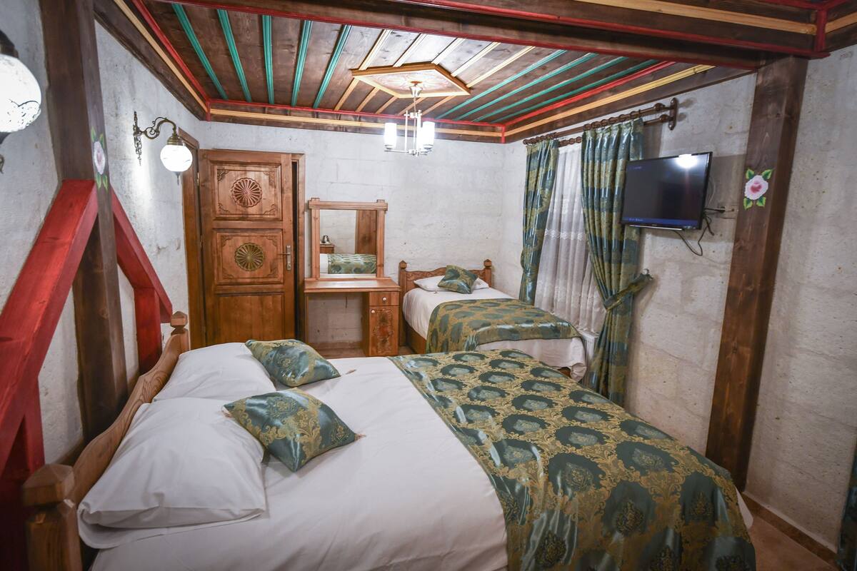 A room with two beds, a wooden wardrobe, a desk, a television, and a ceiling with exposed beams.