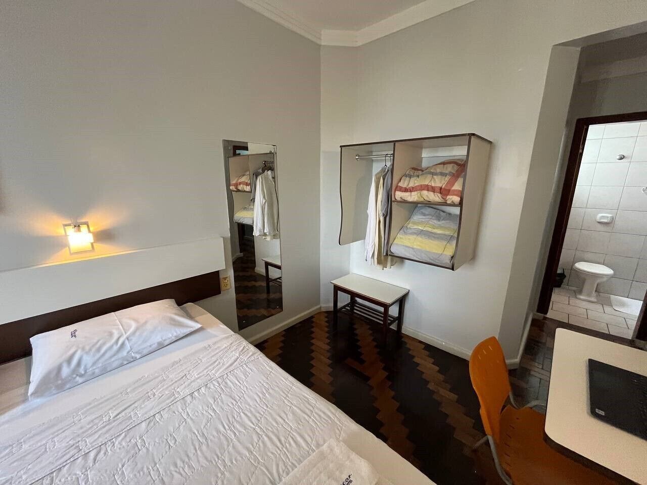 Quarto de hotel com cama, escrivaninha, cadeira, espelho e banheiro visível por uma porta aberta.