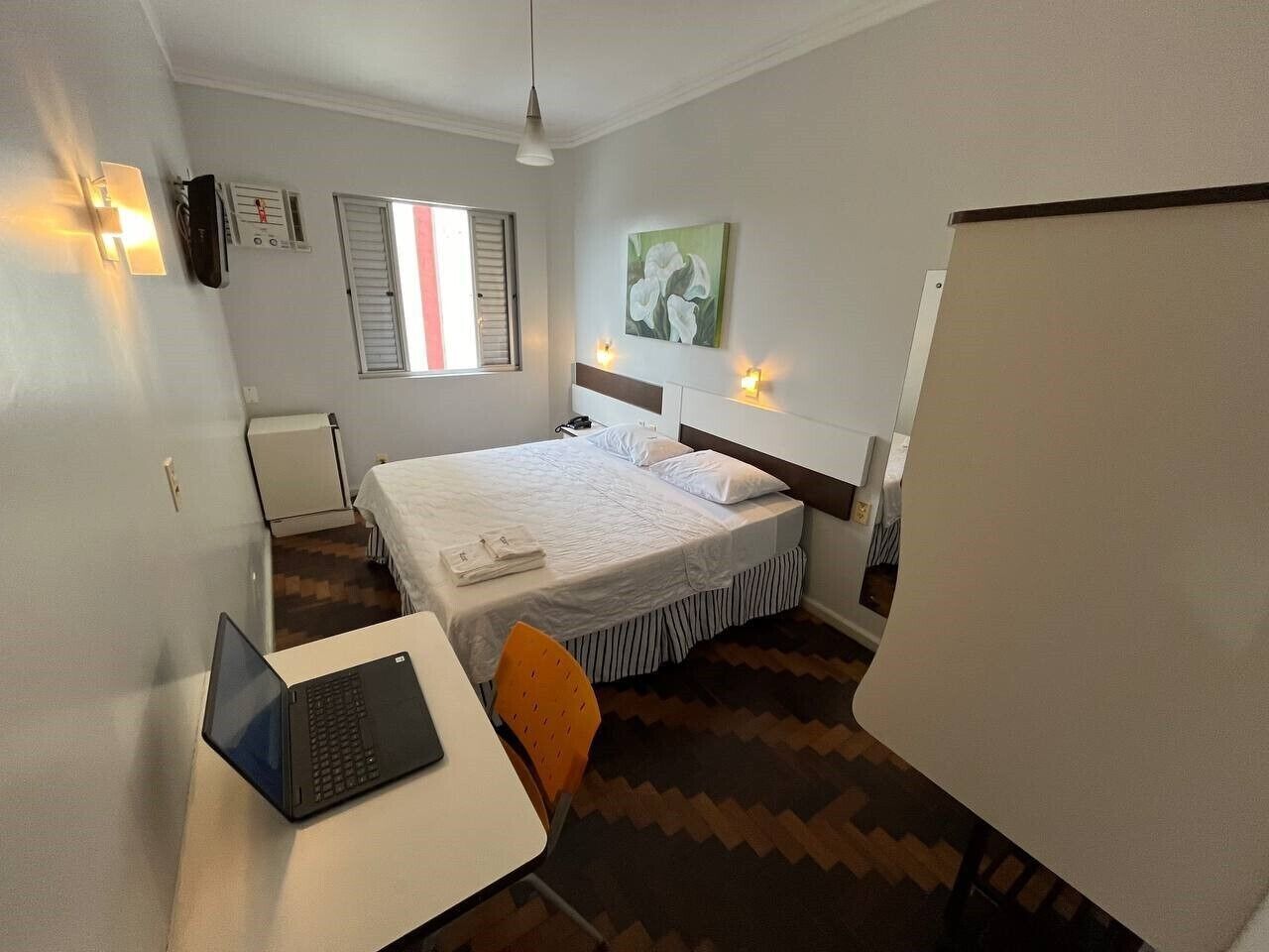 A hotel room with a bed, a desk with a laptop, a chair, a radiator, a window with shutters, and a painting on the wall.