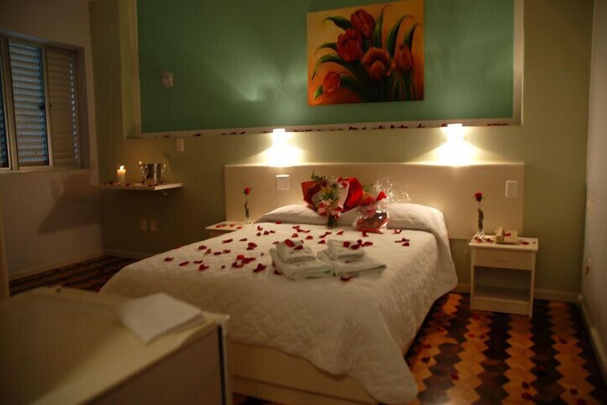 A hotel room with a bed, a nightstand, a bedside table, a painting, and a window with blinds.