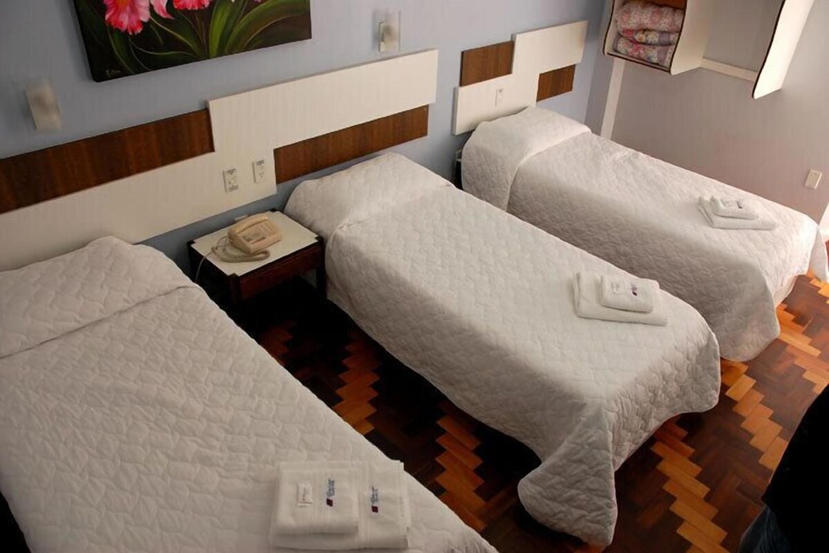 A hotel room with three beds, each with a white quilted cover and a folded towel. There is a wooden headboard with a light switch and a wall-mounted telephone.