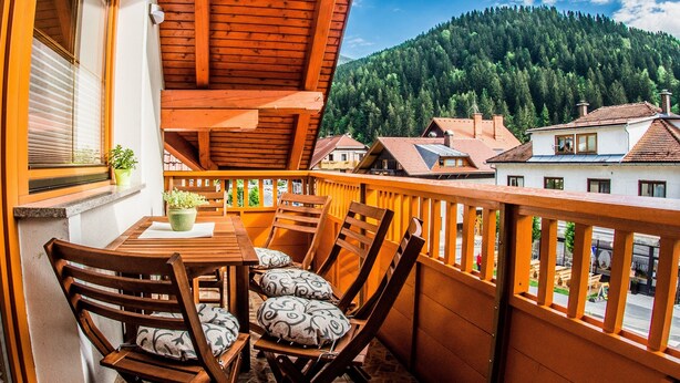 A wooden balcony with a table and chairs, a window with blinds, and a view of a forested hill. - Apartments BBI (Luce)