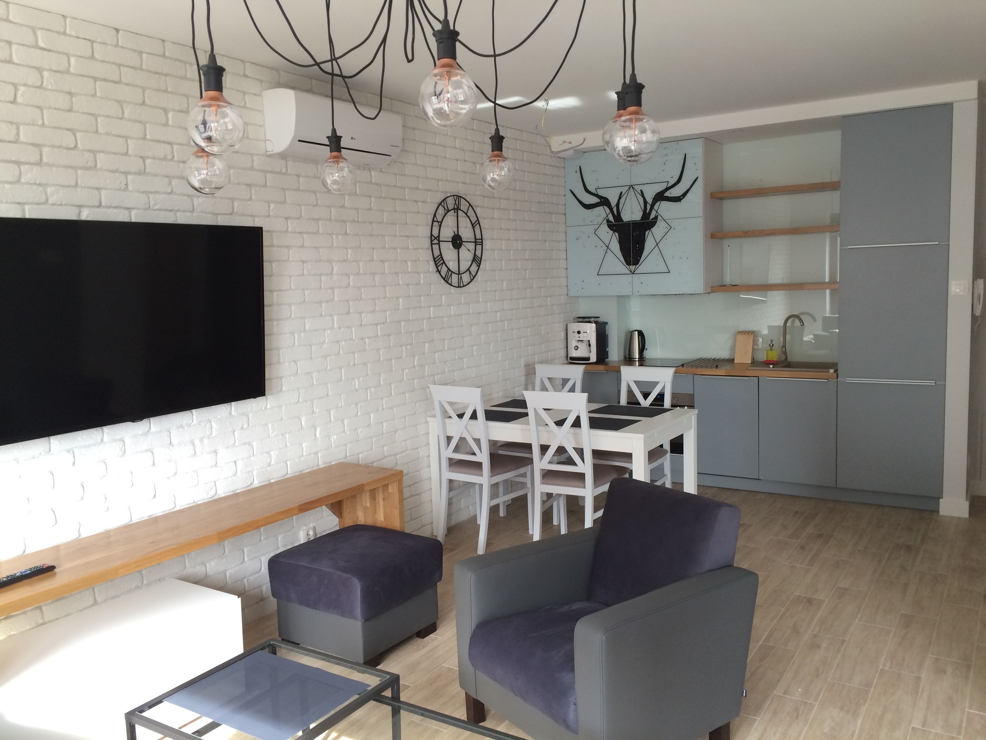 Een moderne woonkamer met een flatscreen televisie, een eethoek met een houten tafel en witte stoelen, en een keuken met grijze kastjes en een blauwe achterwand.