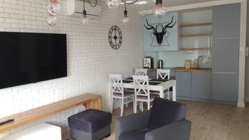 Een moderne woonkamer met een flatscreen televisie, een eethoek met een houten tafel en witte stoelen, en een keuken met grijze kastjes en een blauwe achterwand.