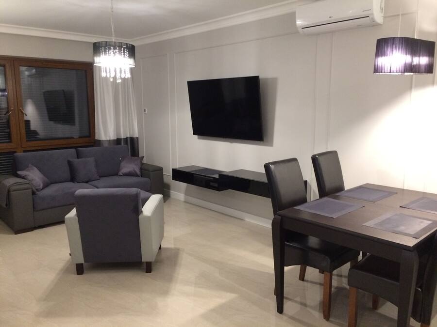 Ein modernes Wohnzimmer mit einem grauen Sofa, einem weißen Hocker, einem Esstisch mit Stühlen, einem flachbildschirmfernseher an der Wand und einer Kronleuchter.