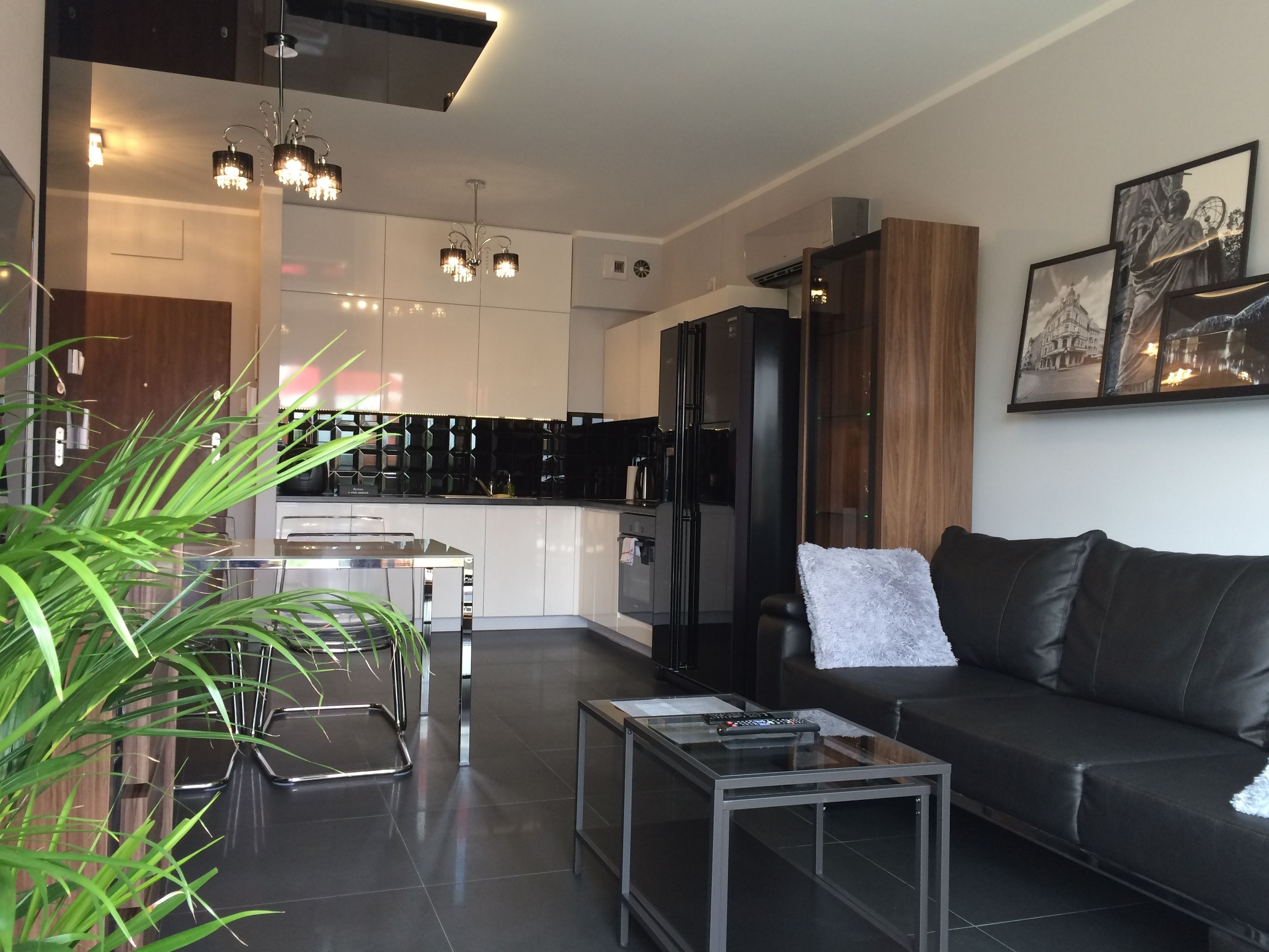A modern living room with a black leather sofa, a glass coffee table, and a dining area with a glass table and chairs. There is a kitchen in the background with black cabinets and a refrigerator.