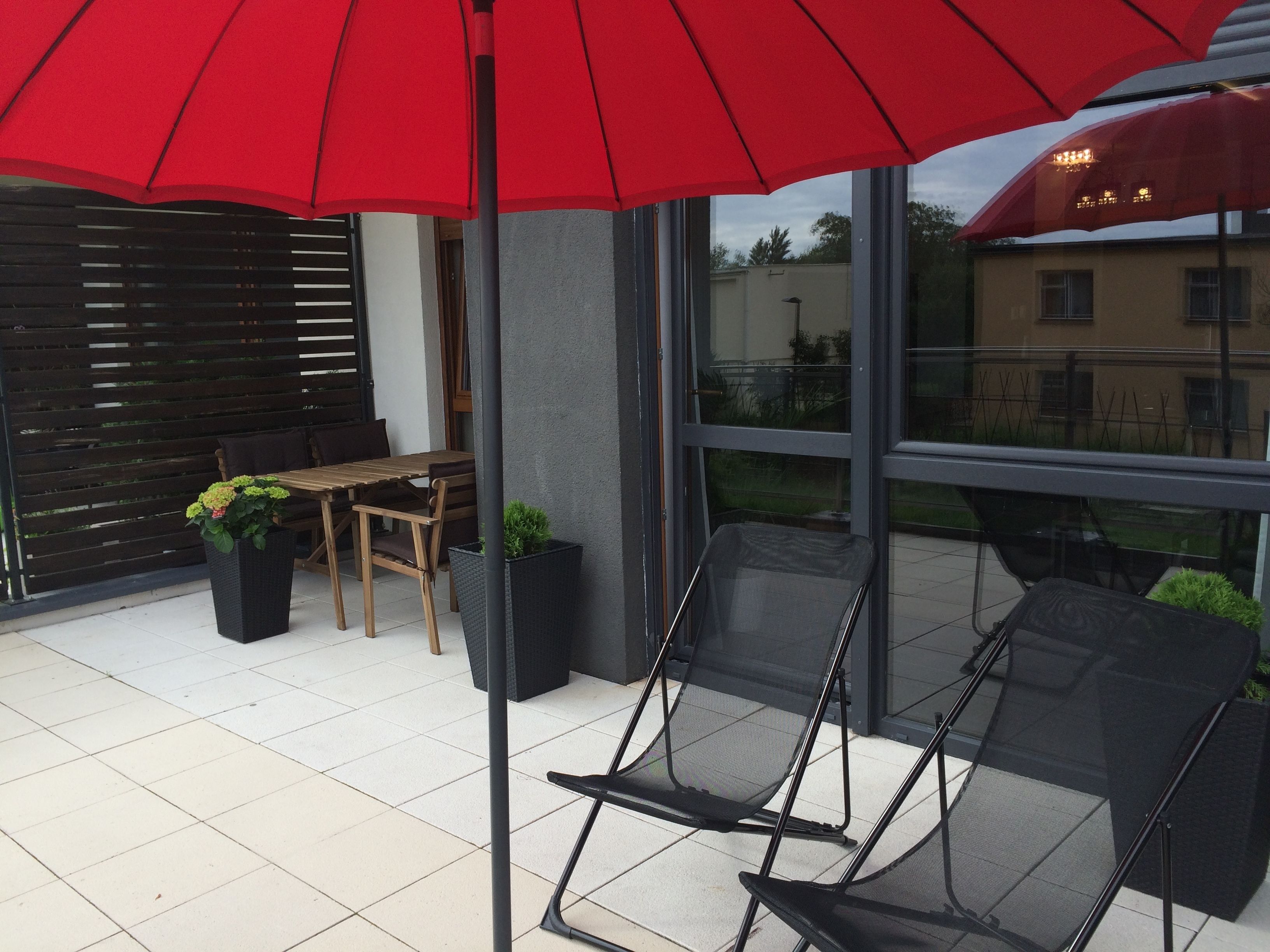 A modern outdoor seating area with a red umbrella, a wooden table, and black chairs.