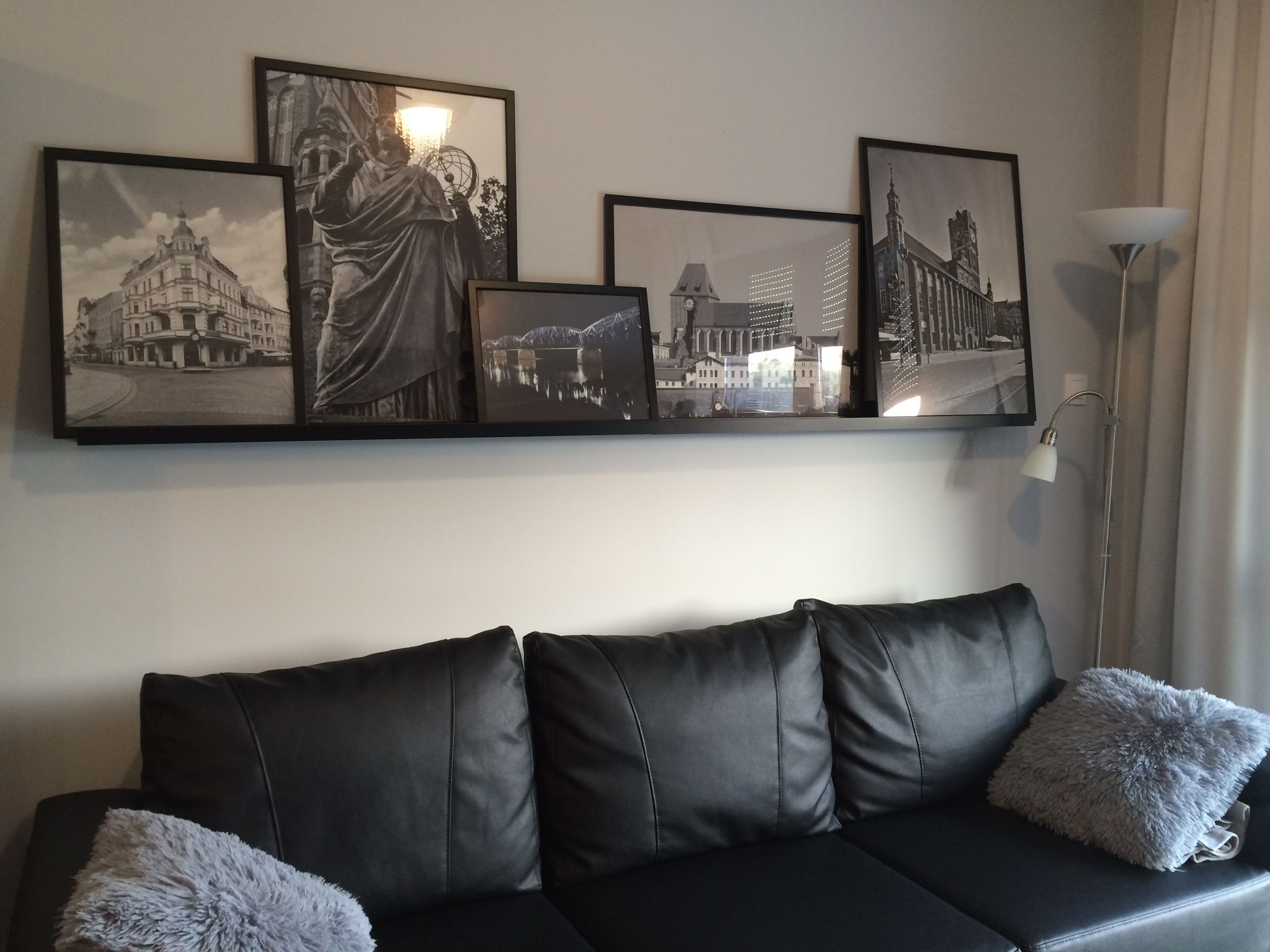 A black leather sofa with fluffy pillows, a floor lamp, and a wall with framed black and white cityscape photographs.