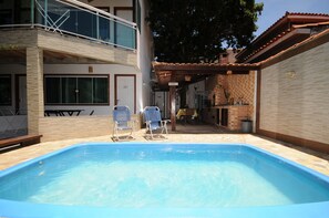 Piscina com duas espreguiçadeiras, um terraço coberto e vista para um prédio com varanda.
