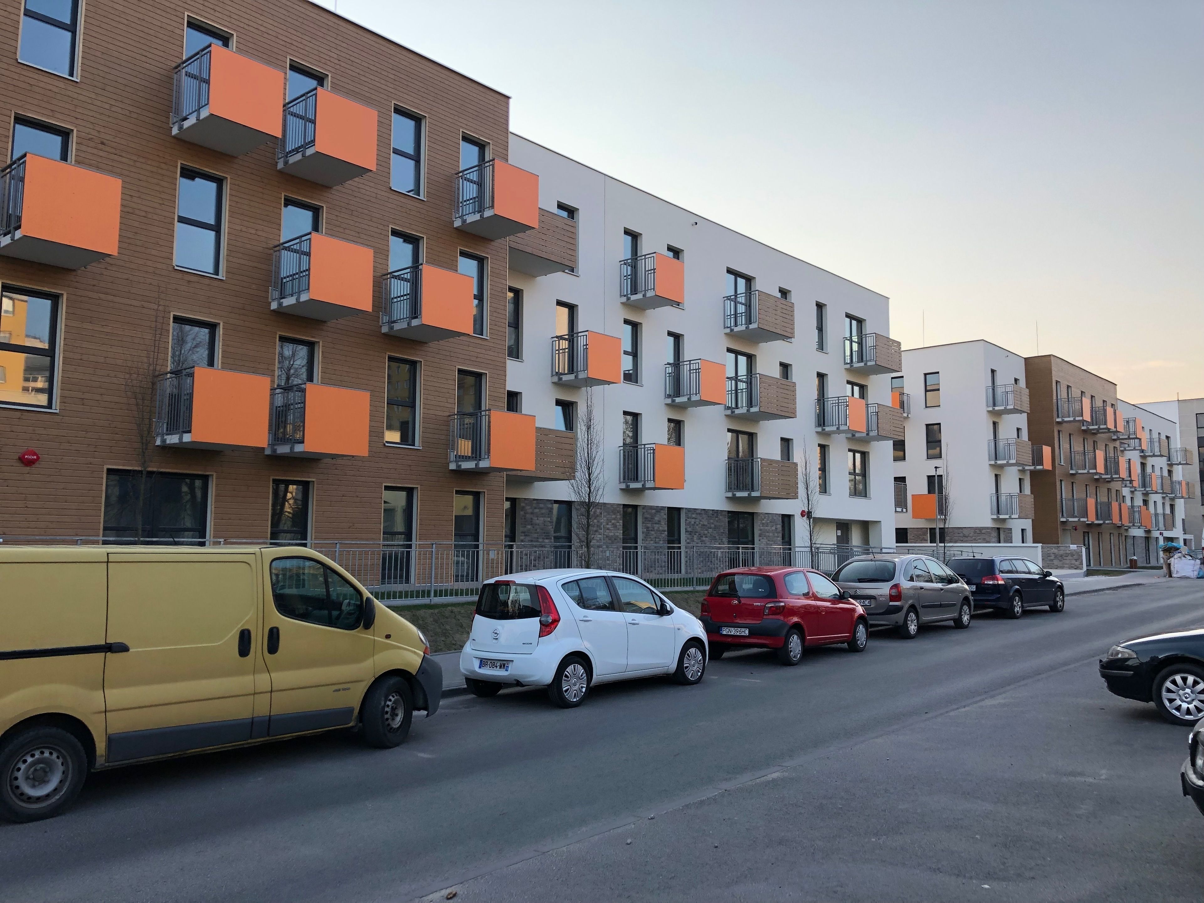 Una hilera de edificios de apartamentos modernos con balcones, una furgoneta amarilla y varios coches estacionados a lo largo de la calle.