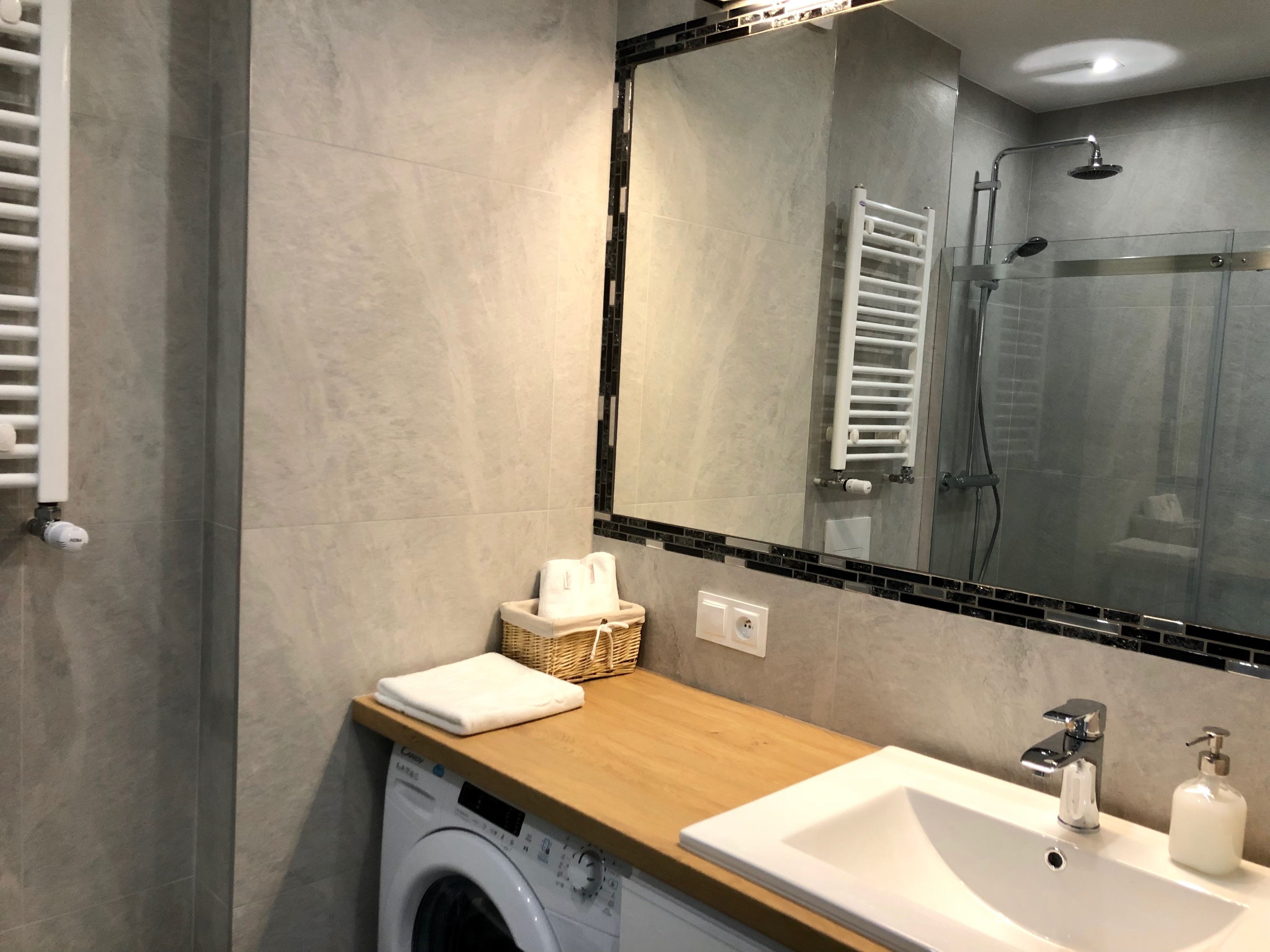A modern bathroom with a built-in washing machine, a wooden countertop, a sink with a soap dispenser, and a glass shower enclosure.