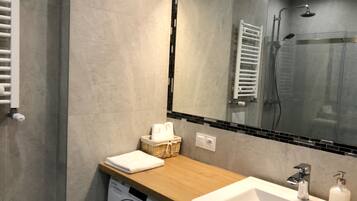 A modern bathroom with a built-in washing machine, a wooden countertop, a sink with a soap dispenser, and a glass shower enclosure.