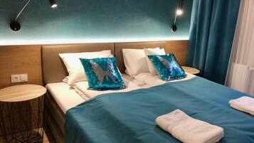 A neatly made bed with a teal bedspread and two decorative pillows, a small round side table, and a wall-mounted lamp.