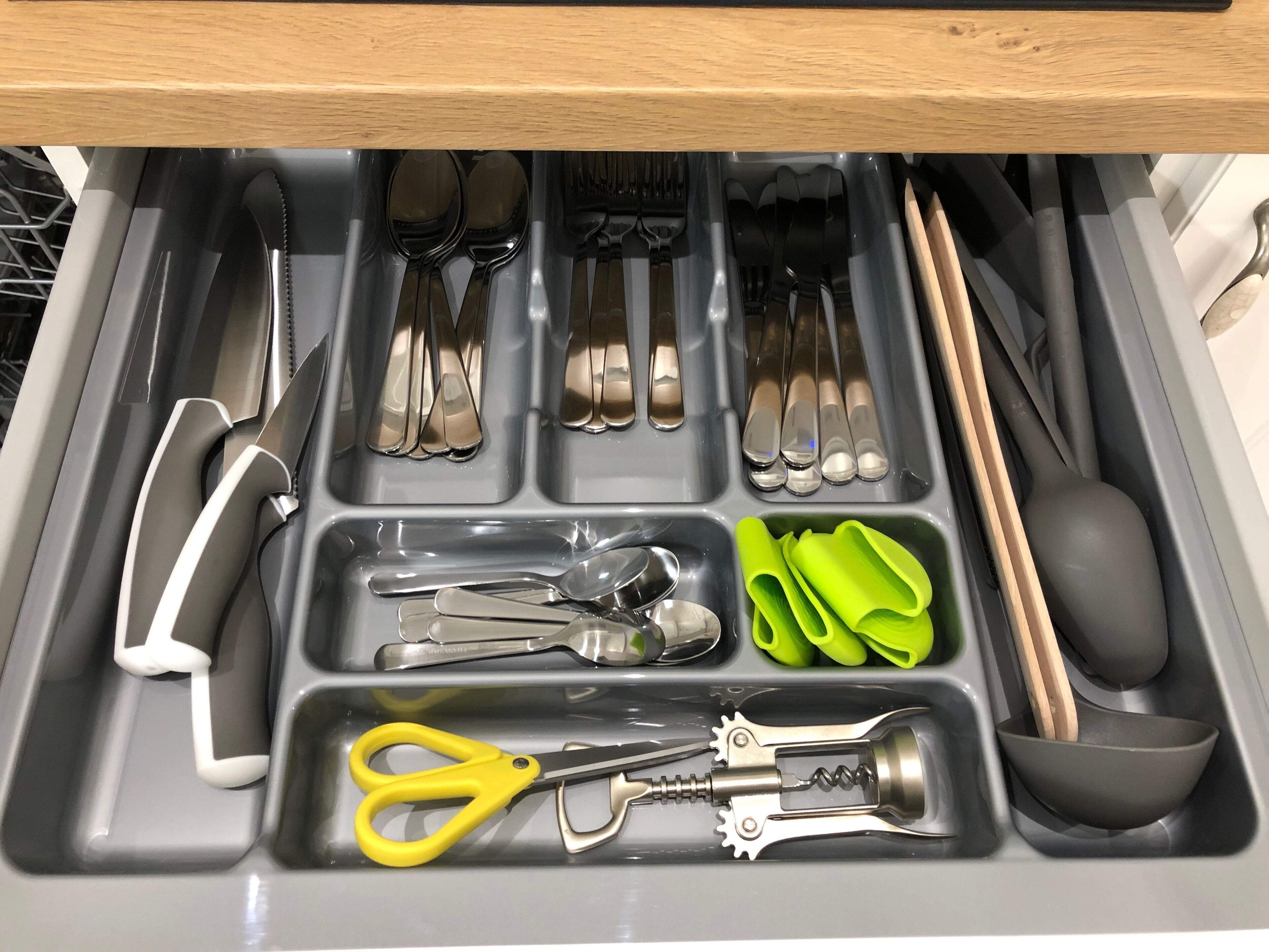 A drawer with various utensils and tools including scissors, a corkscrew, and green silicone grips.