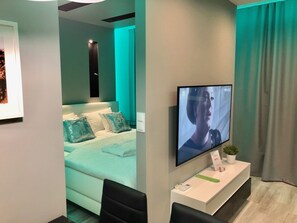 A modern hotel room with a bed, a TV mounted on the wall, a small desk with a plant, and a window with curtains. - OldTown deLuxe (Torun)