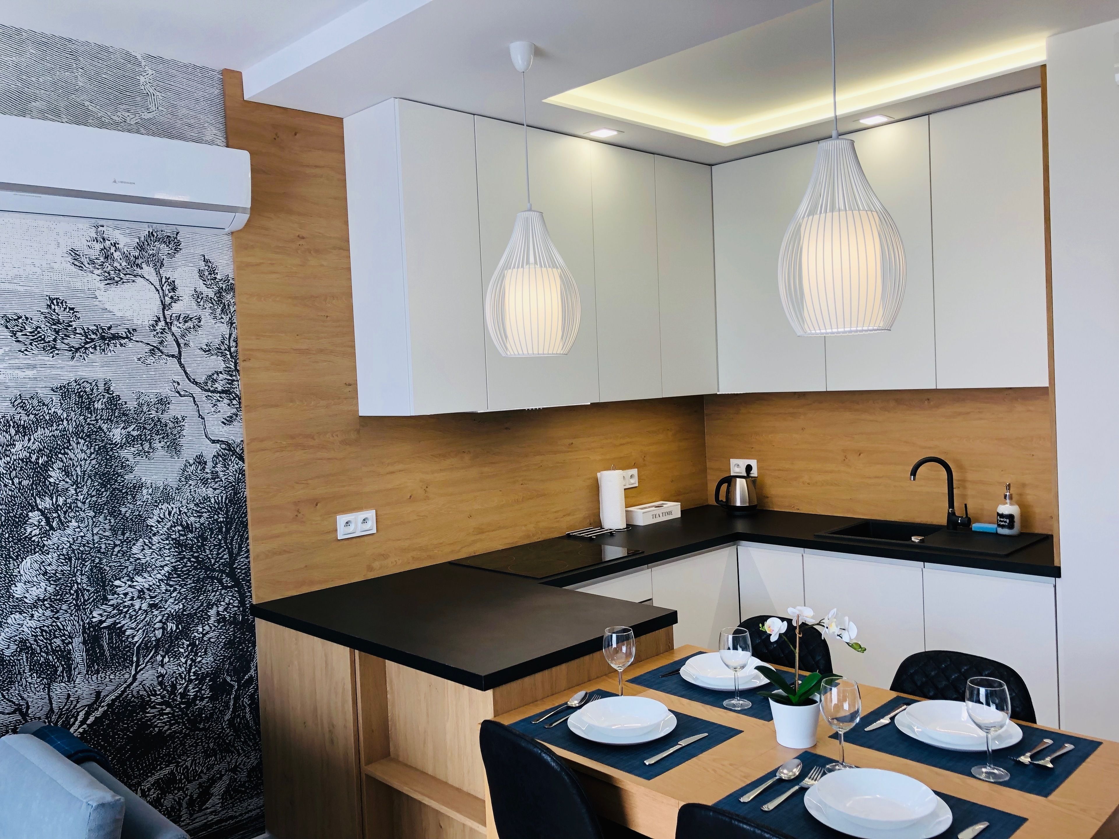 A modern kitchen with white cabinets, a black countertop, and pendant lights. There is a dining table set with plates, cutlery, and a vase with flowers.