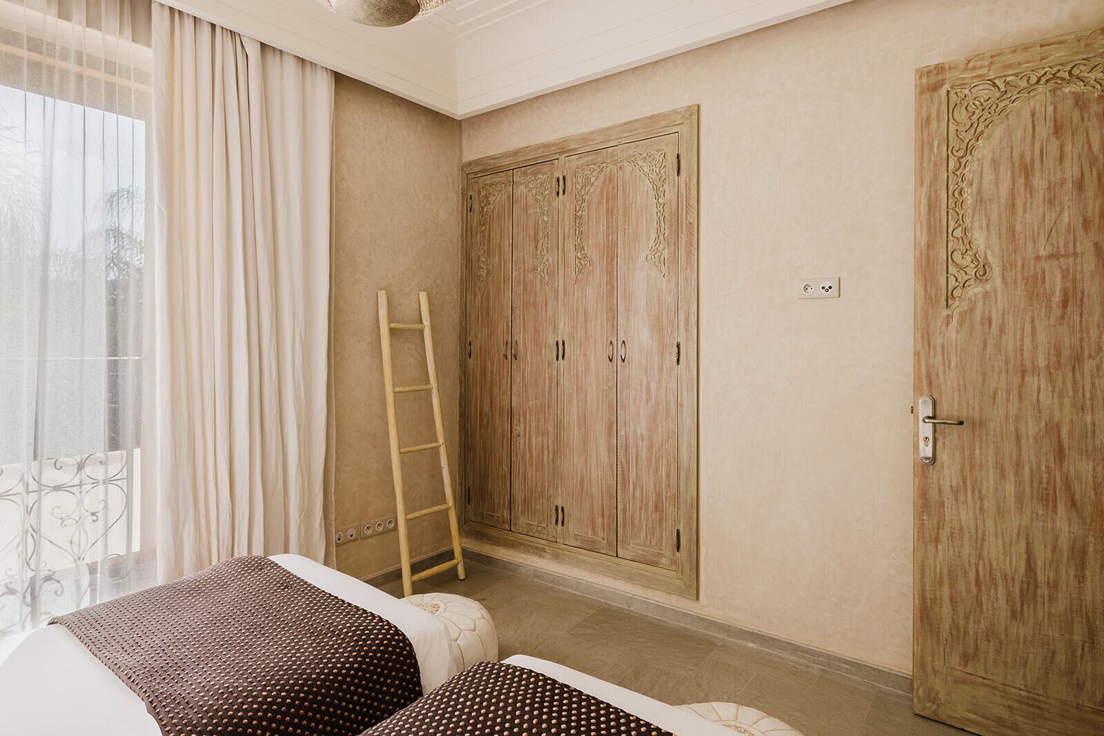 Ein Schlafzimmer mit großem Kleiderschrank, einer Hockerleiter und einem Bett mit gemusterten Betttextilien.