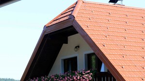 A two-story building with a sloped roof, a balcony with flower boxes, and a view of a forested mountain. - Guest House Koprivnik (Zrece)