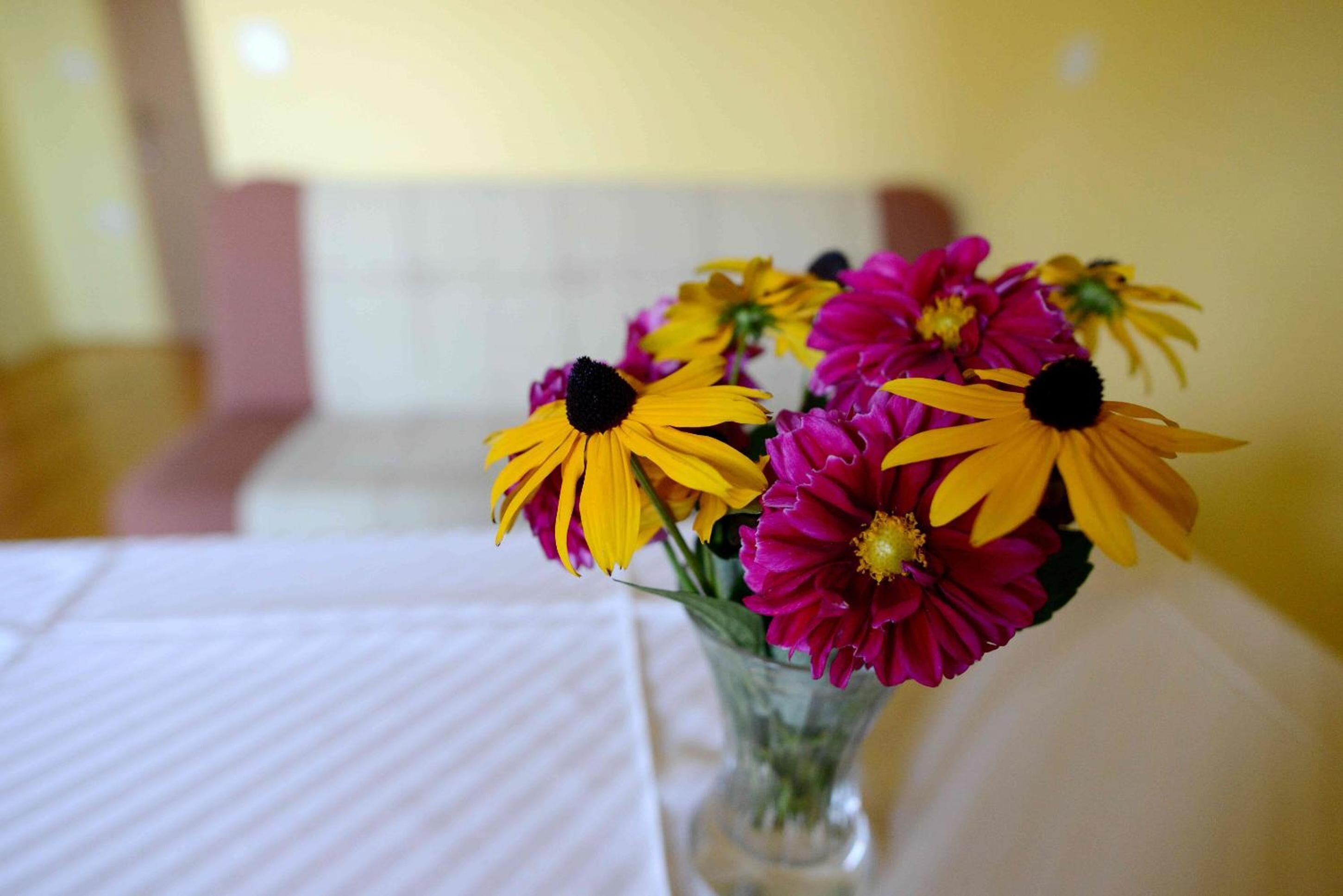 A clear view of a table with a white tablecloth, a clear glass vase, and a bouquet of flowers with yellow and pink petals.