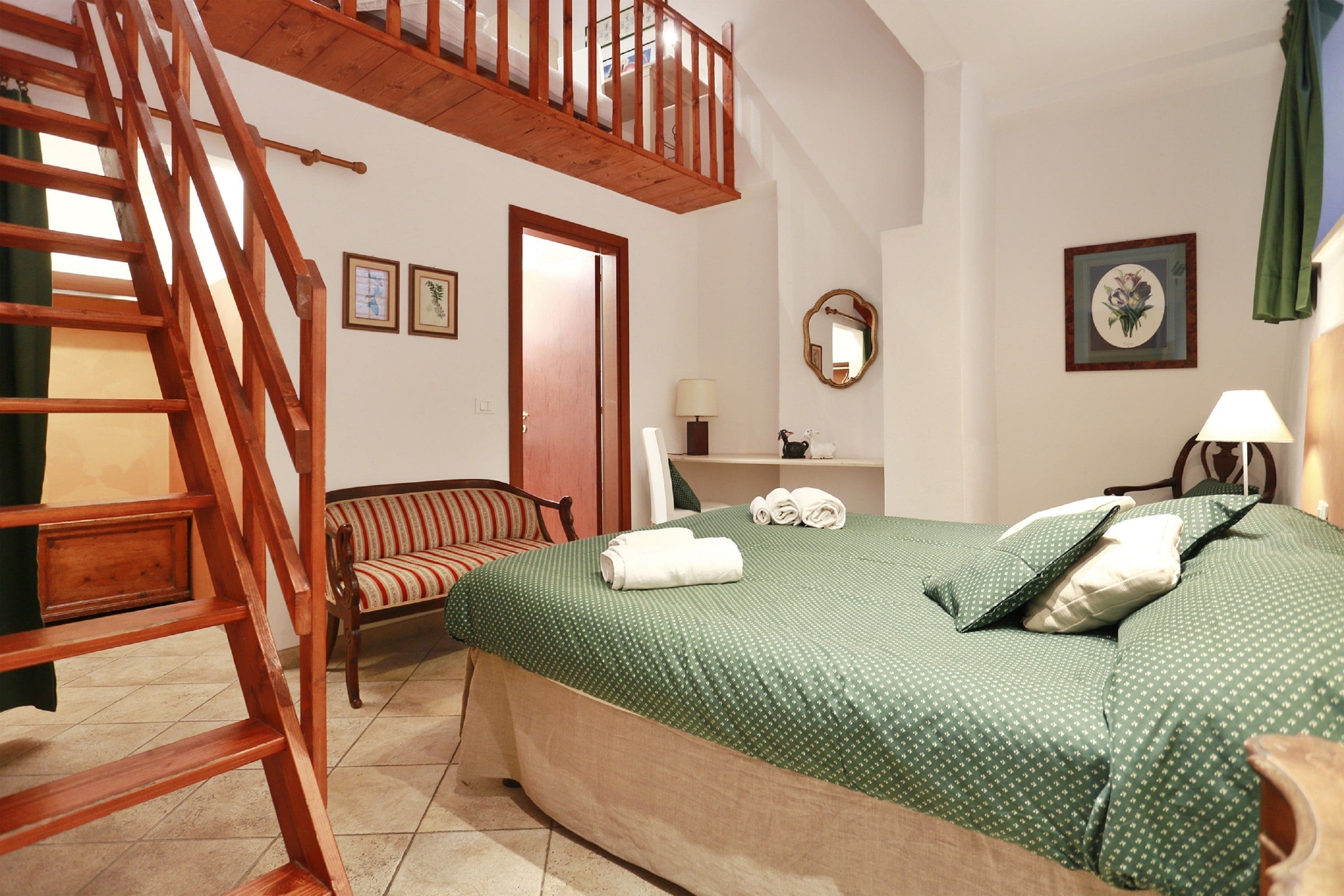 Una camera da letto con una scala in legno, un letto con lenzuola verdi, una sedia, un comodino con una lampada e uno specchio a parete.