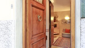 A wooden door with a brass handle, leading into a room with a patterned rug, a sofa, and a wall-mounted shelf with decorative items. - Ghibellina Apartment (Florence)