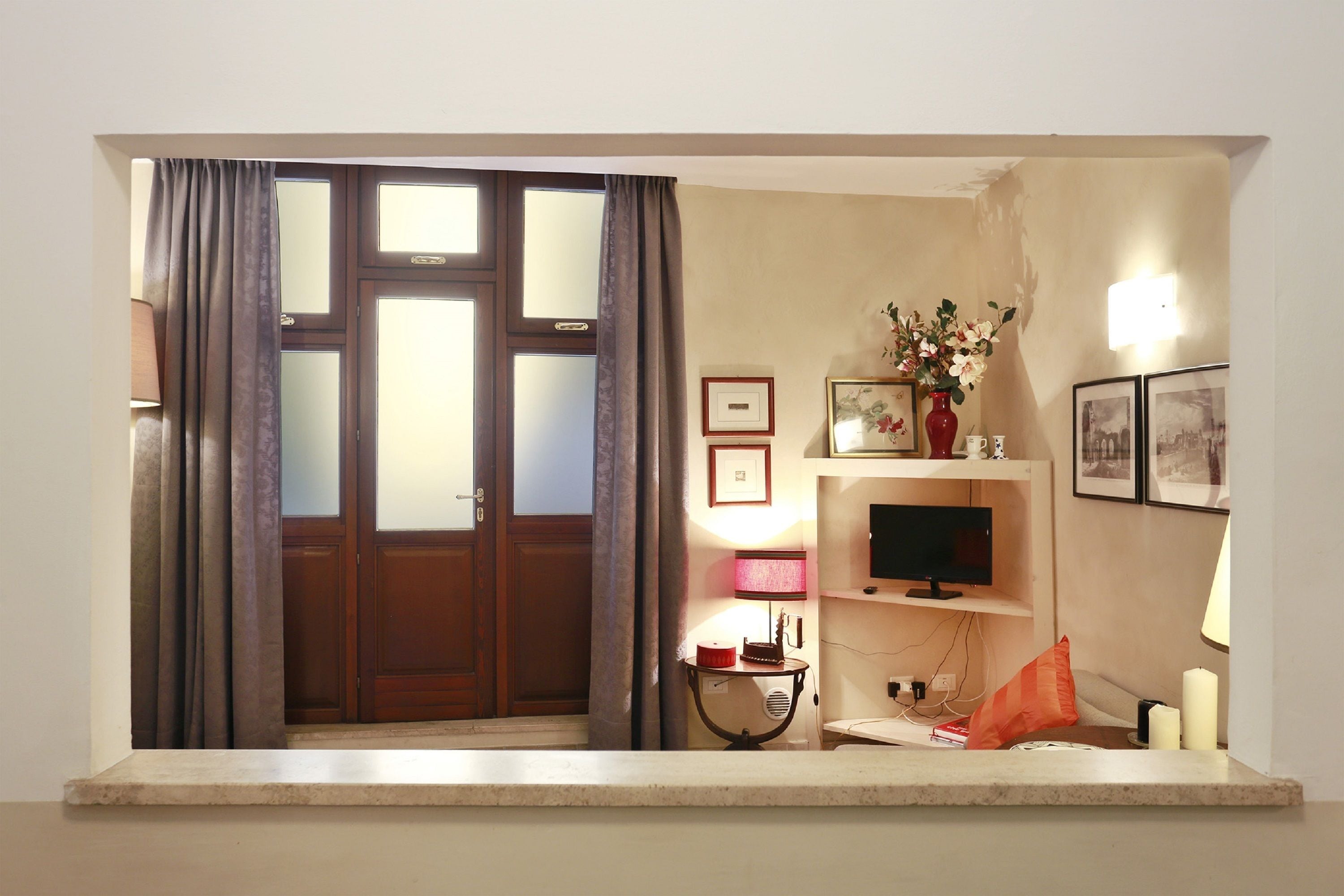 A room with a wooden door, a television on a stand, a small table with a lamp, and a shelf with framed pictures.