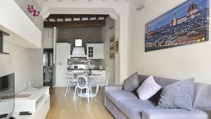 Una sala de estar moderna con un sofá gris, un comedor con sillas blancas y una cocina con armarios blancos.