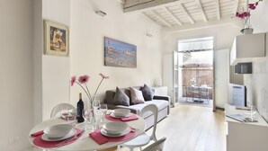 Un comedor con una mesa blanca para cuatro personas, un cuadro de un paisaje urbano y un sofá con cojines al fondo.