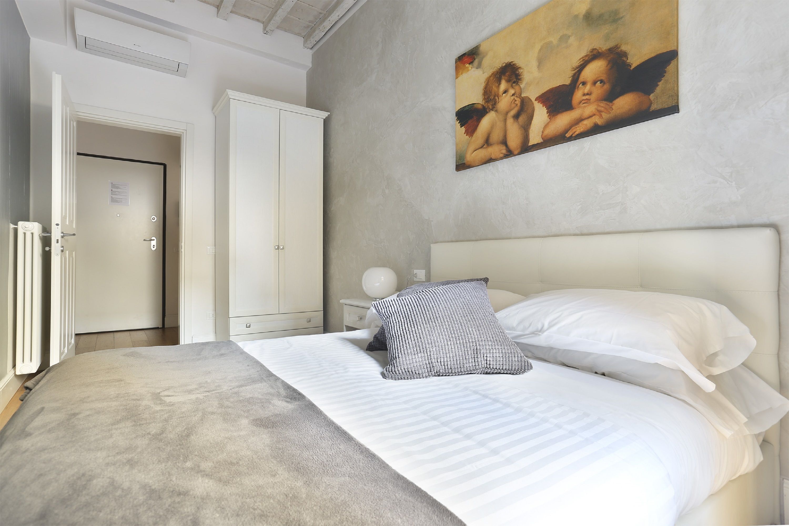 A bedroom with a large bed, white bedding, a nightstand with a lamp, a wardrobe, and a painting on the wall.