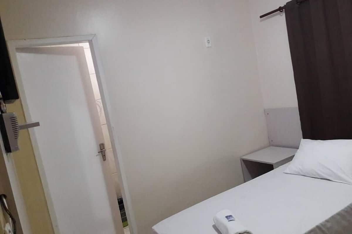 A small, simple hotel room with a bed, a nightstand, a door, and a curtain.