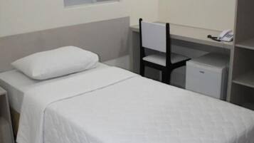 Una cama individual con colchón y almohada blancos, una silla, un escritorio pequeño con silla, y un teléfono en un estante.