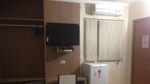 A small room with a television mounted on the wall, a refrigerator, a chair, and a window with curtains. - Hotel Express (Varzea Grande)