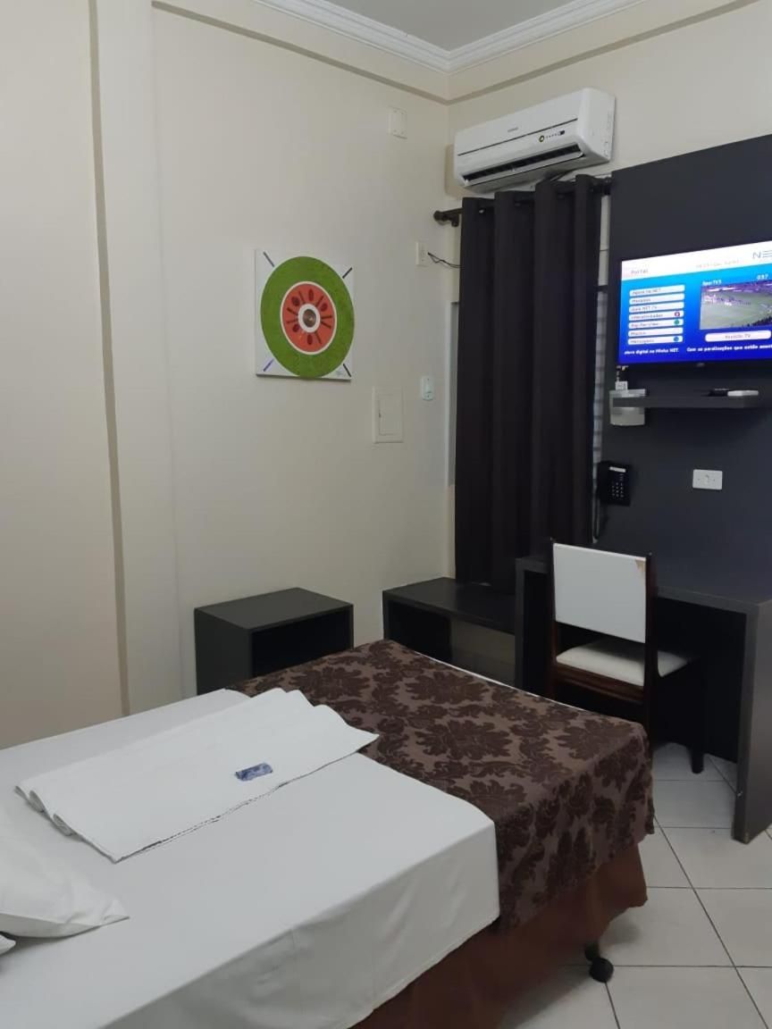 A hotel room with a bed, a desk, a chair, an air conditioning unit, a television, and a wall-mounted target.