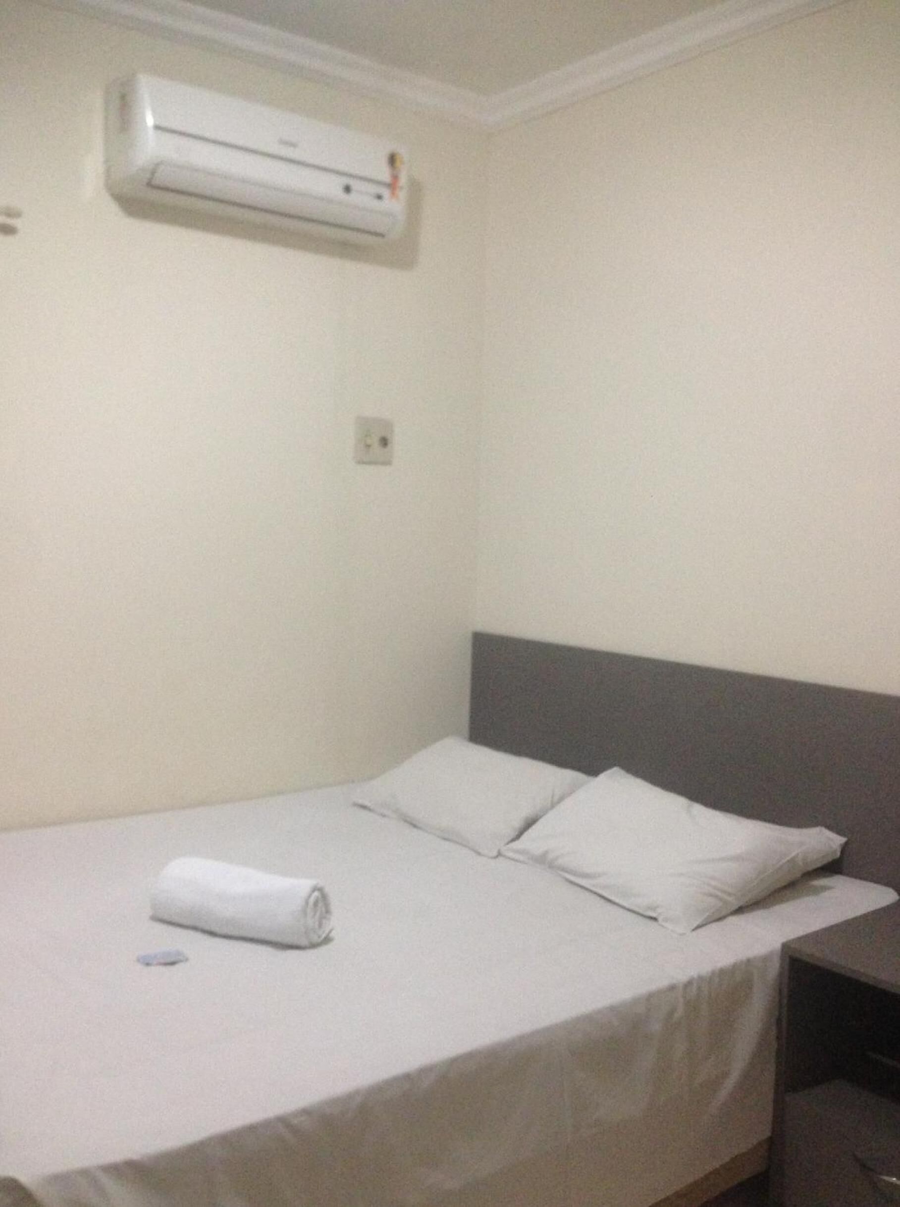 A single bed with a grey headboard and white bedding in a room with a white wall and an air conditioning unit mounted above it.
