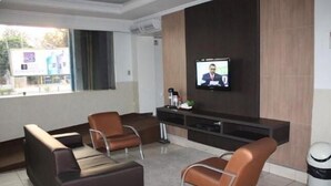 A modern waiting area with leather sofas, a flat-screen TV mounted on the wall, and a trash bin. - Hotel Express (Varzea Grande)