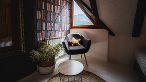 A cozy reading nook with a bookshelf, a chair, and a small table in a room with sloped ceilings and a window. - Apartments Wanderlust (Bovec)