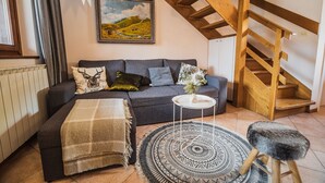 A cozy living room with a grey sofa, a small white side table, a patterned rug, a wooden stool, and a staircase with exposed beams. - Apartments Wanderlust (Bovec)