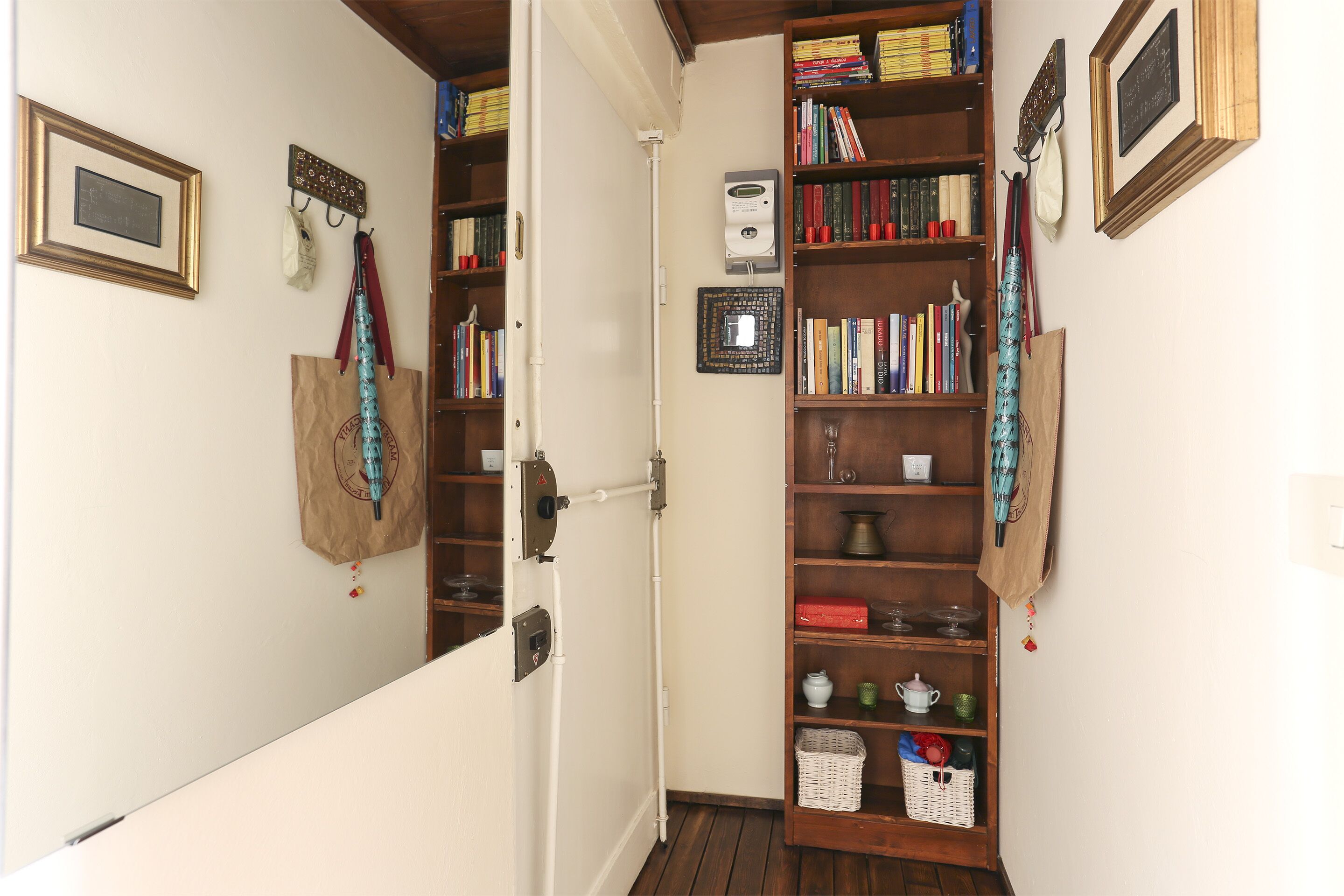 Un couloir étroit avec une bibliothèque en bois remplie de livres et d’objets décoratifs, une porte blanche et un petit panier blanc posé sur le sol.