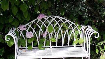 A white metal bench with a lattice backrest, surrounded by lush green foliage and purple flowers.