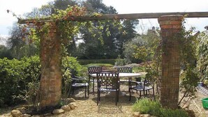 Een tuinpatio met een pergola, stenen pilaren en een ronde tafel met stoelen.