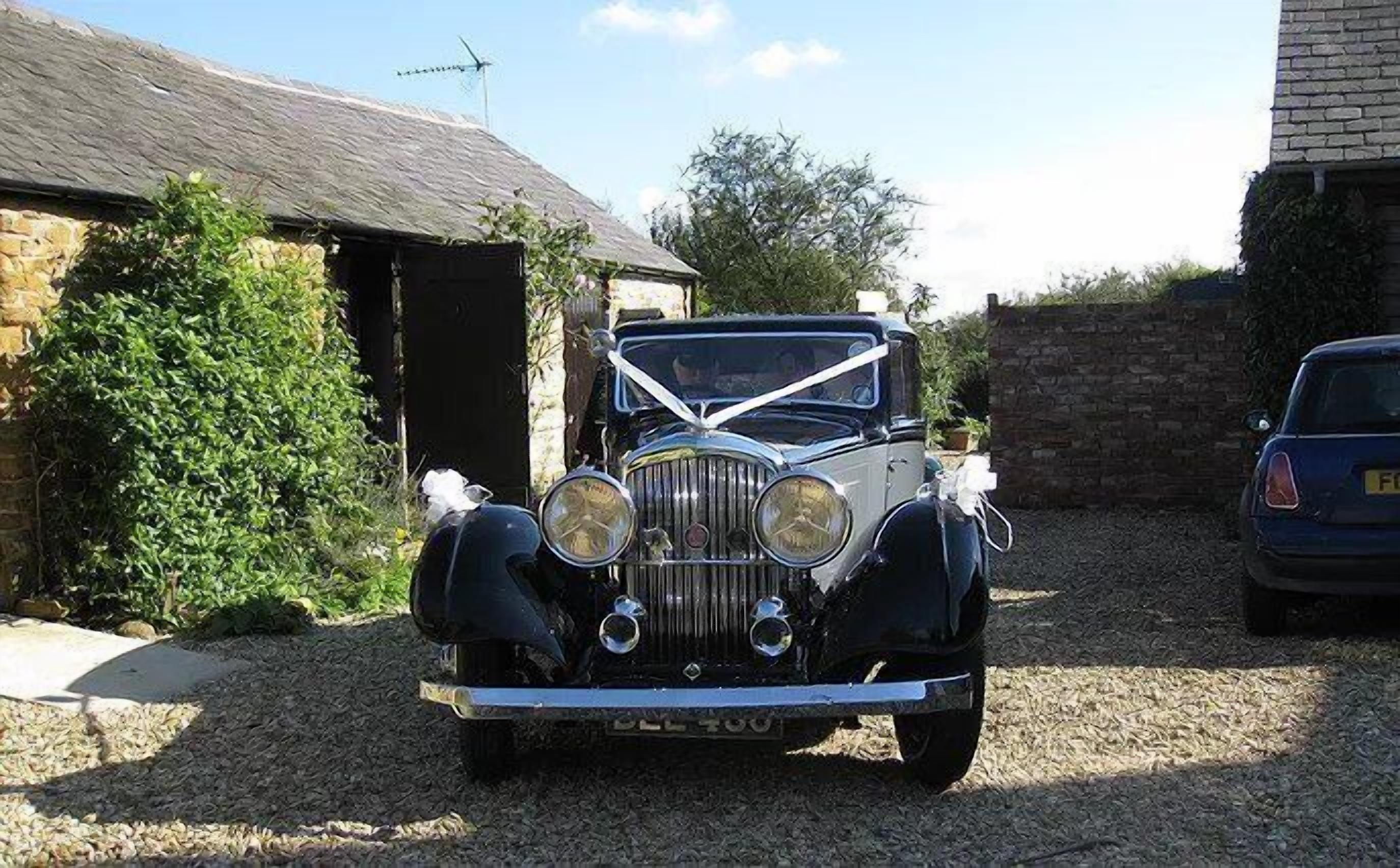 A vintage car parked in a driveway with a stone building and a hedge in the background.