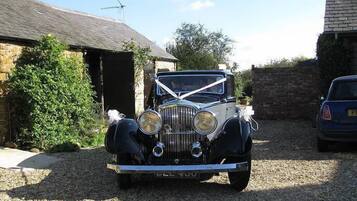 A vintage car parked in a driveway with a stone building and a hedge in the background.