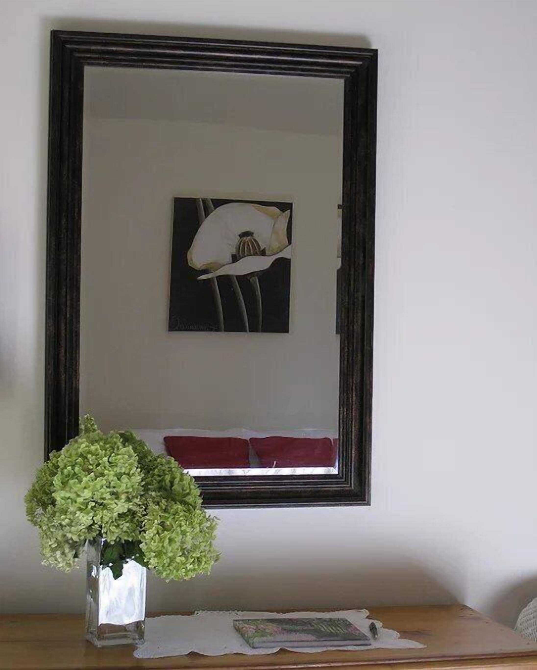 A framed artwork of a white flower on a wall, a vase with green foliage on a table, and a red cushion on a bench.