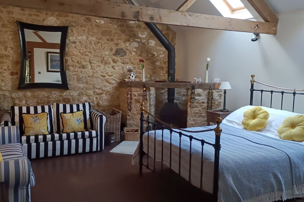 Een knus interieur met een stenen muur, een bed met gele kussens, een gestreept bankje en een stoel met metalen frame.