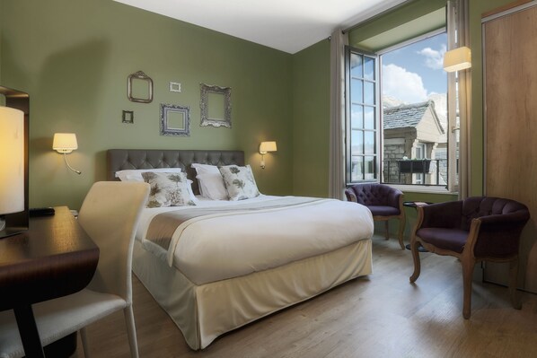 A bedroom with a large bed, two chairs, a desk, and a window with a view of a building. - Hôtel Joyet de Maubec (Uzerche)