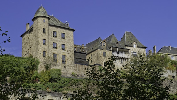 A large stone building with multiple towers and a balcony, surrounded by greenery. - Hôtel Joyet de Maubec (Uzerche)