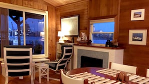 A cozy cabin interior with wooden walls, a fireplace, a dining table with placemats, and a view of the outside through a large window.