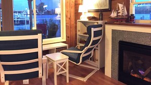A cozy room with wooden walls, a fireplace, a window with a view of a dock, and a blue chair.