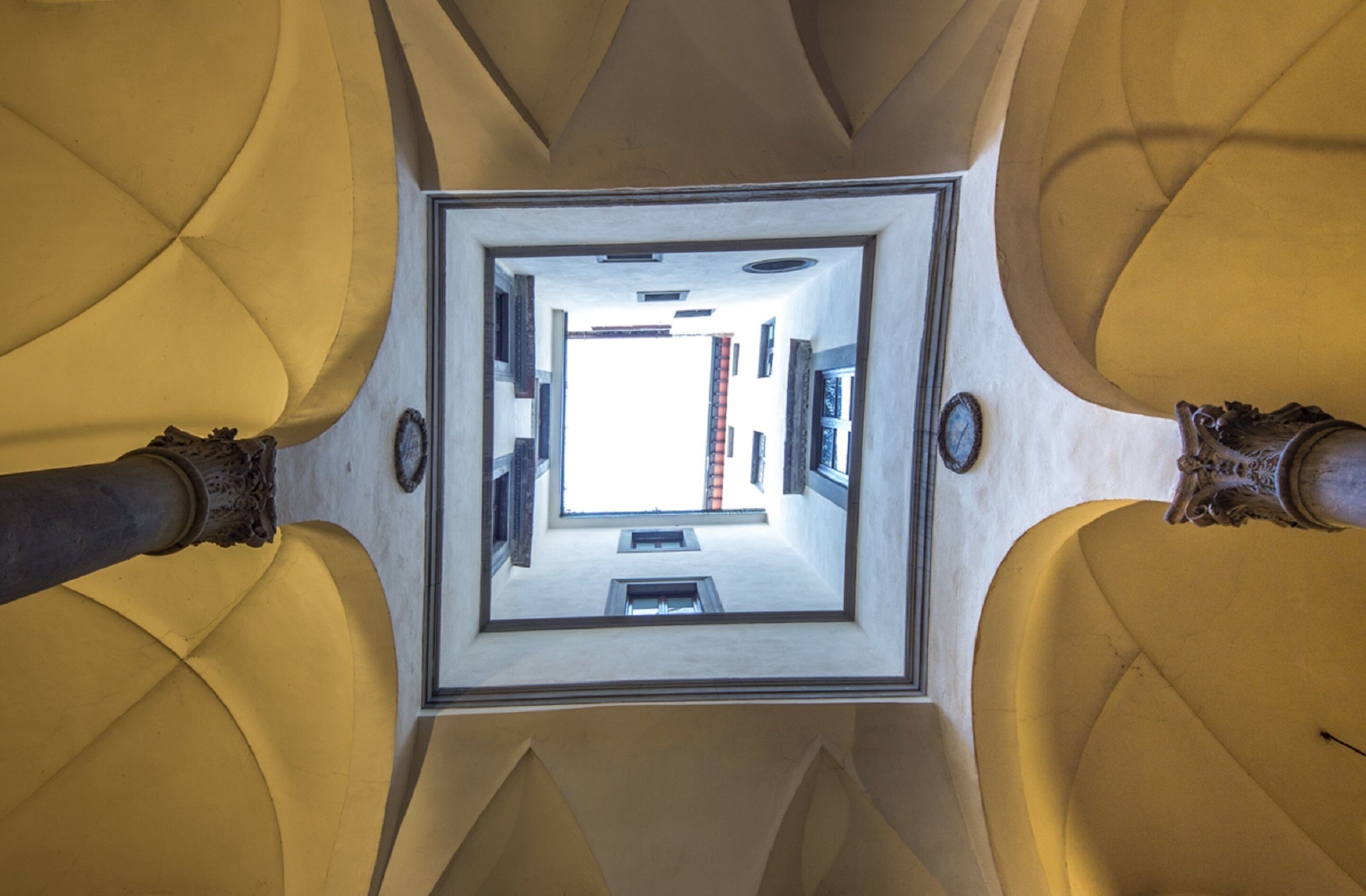 A view from a high ceiling looking down through a square opening, revealing a room with a bed, a window, and a door.